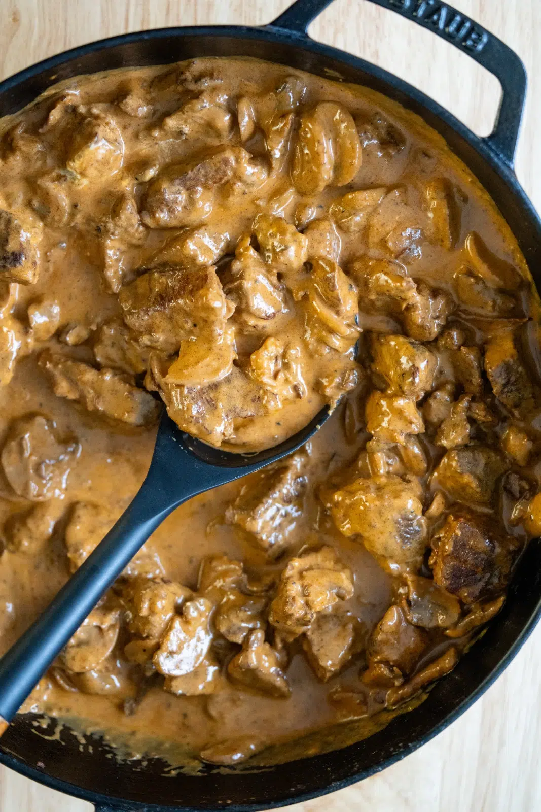 Overhead view of a cast iron pan filled with creamy slow-cooked beef stroganoff, featuring tender chunks of beef and sliced mushrooms coated in a rich, velvety brown sauce, with a black serving spoon lifting a portion from the pan.