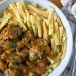 Top view of a white speckled bowl filled with slow-cooked beef stroganoff and pasta, showcasing tender beef in a rich creamy sauce alongside twisted pasta noodles, garnished with chopped parsley and served on a wooden table with a checkered napkin and silver cutlery.