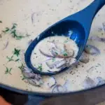 Close-up of creamy mushroom sauce with sliced mushrooms and fresh thyme in a black ladle over a skillet, ready for serving.