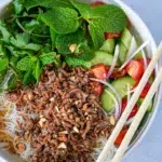 Bowl of Thai beef and noodle salad featuring seasoned ground beef, topped with sliced cucumber, rice noodles, fresh herbs, and onions, with chopsticks on the side