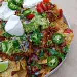 A bowl of crispy nachos loaded with pulled chicken, avocado, jalapeños, sour cream, tomatoes, and fresh cilantro