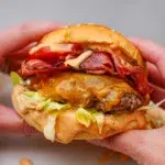 Two hands gripping a beef and bacon burger, ready for a big bite.