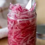 Jar of homemade pickled red onions with a fork, showcasing vibrant pink color and crisp texture.