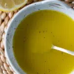 Close-up of a simple lemon vinaigrette with black pepper in a white ceramic bowl, next to a halved lemon.
