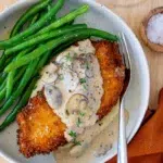 Crispy breaded chicken schnitzel topped with creamy mushroom sauce, served with sautéed green beans and mashed potatoes in a bowl.