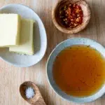 Ingredients for easy hot honey sauce on a wooden surface, including butter, chili flakes, honey, and salt displayed in small bowls and a wooden spoon.