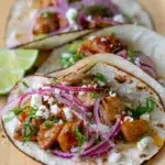 Sticky lime honey glazed chicken tacos topped with pickled red onions, crumbled cheese, and fresh herbs in toasted flour tortillas, served with lime wedges.