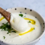 Thick and creamy garlic yoghurt sauce in a saucer bowl, lifted with a wooden spoon