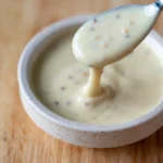 Creamy honey mustard dipping sauce in a small bowl, with a spoon lifting the sauce to show its smooth texture and mustard seeds.