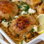 Baked one pan Greek chicken thighs with golden crispy skin, served over risoni pasta with spinach, feta, and lemon slices in a white baking dish.