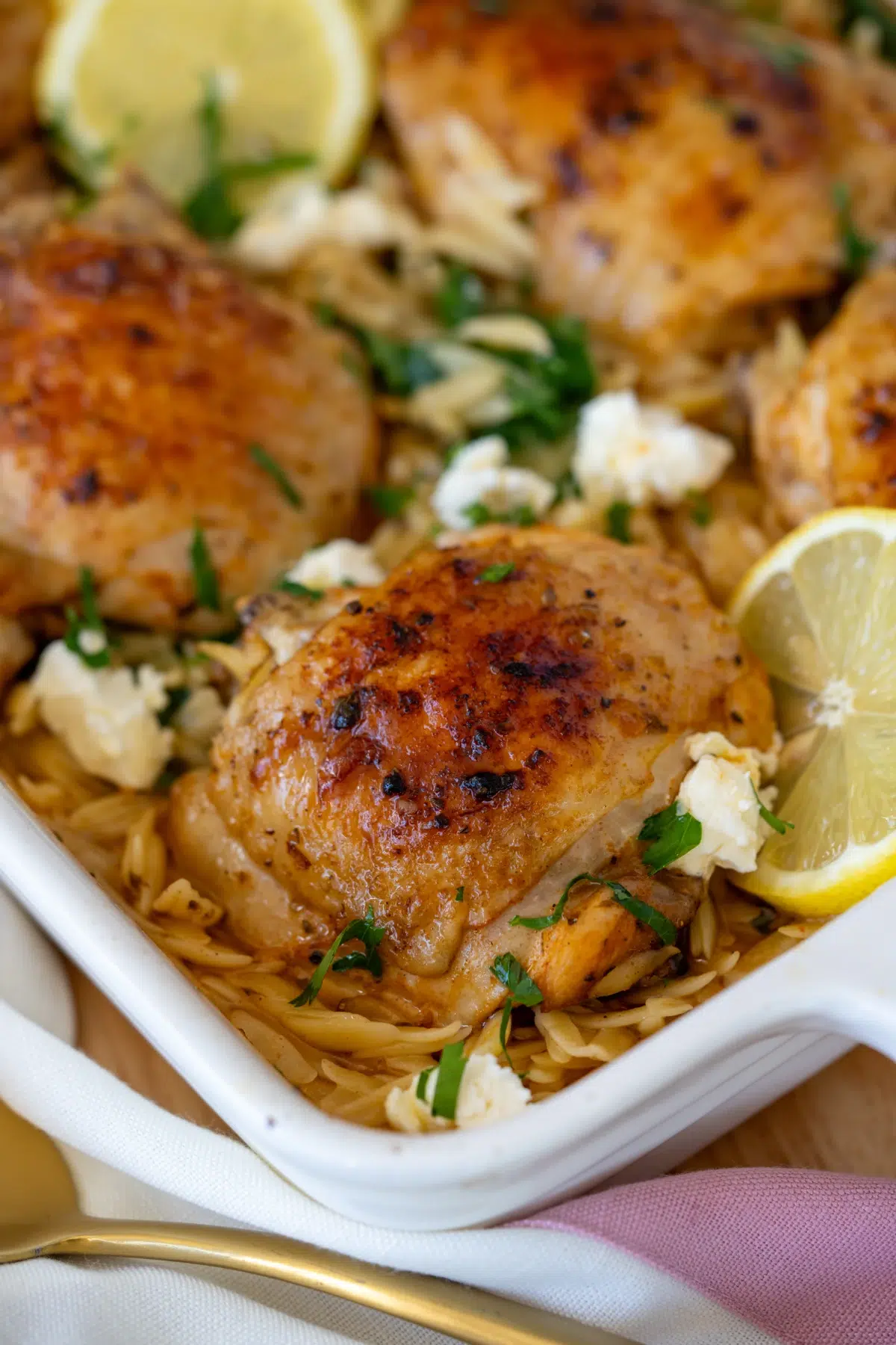 Baked one pan Greek chicken thighs with golden crispy skin, served over risoni pasta with spinach, feta, and lemon slices in a white baking dish.