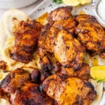Overhead close-up of golden charred garlic lemon chicken thighs piled on flatbread on a white speckled platter with lemon wedges and crispy roasted potatoes