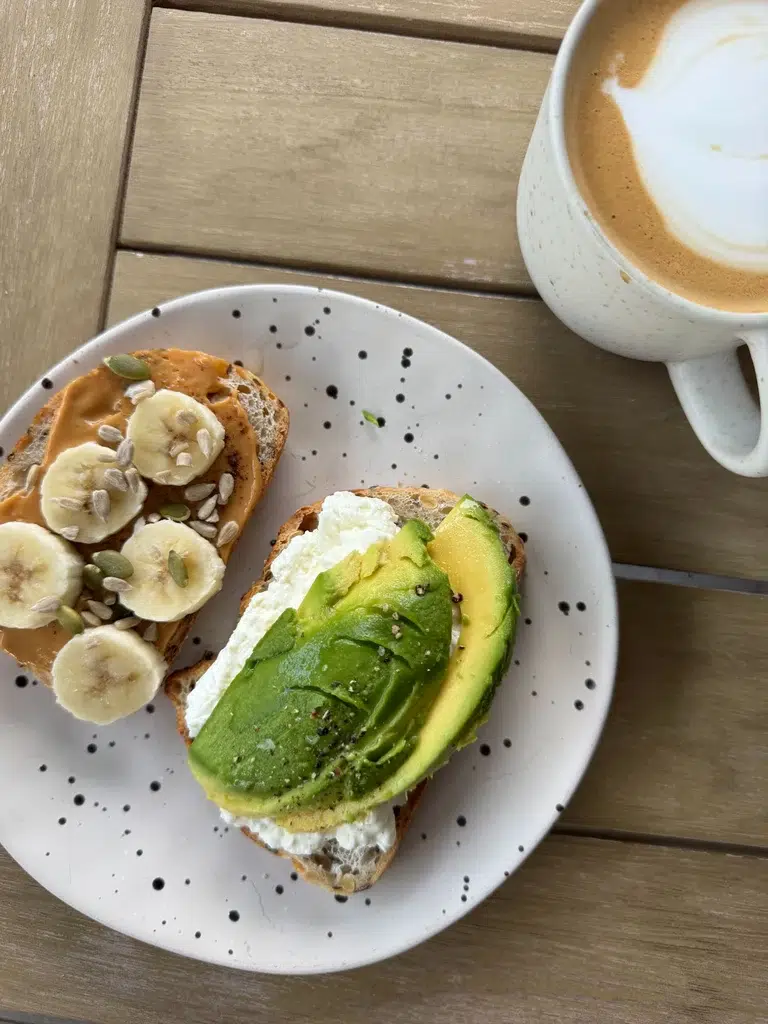Sourdough toast with cottage cheese and avocado, and peanut butter with banana and seeds, beside a coffee