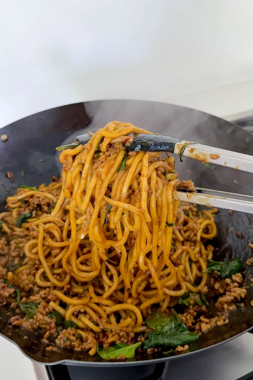 noodles being tossed with tongs in wok