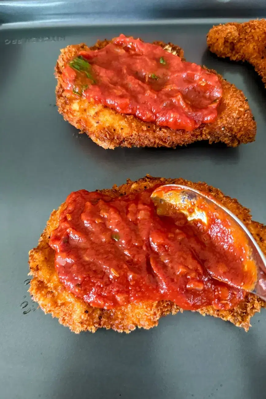 Cooked chicken schnitzels being topped with tomato and basil sauce
