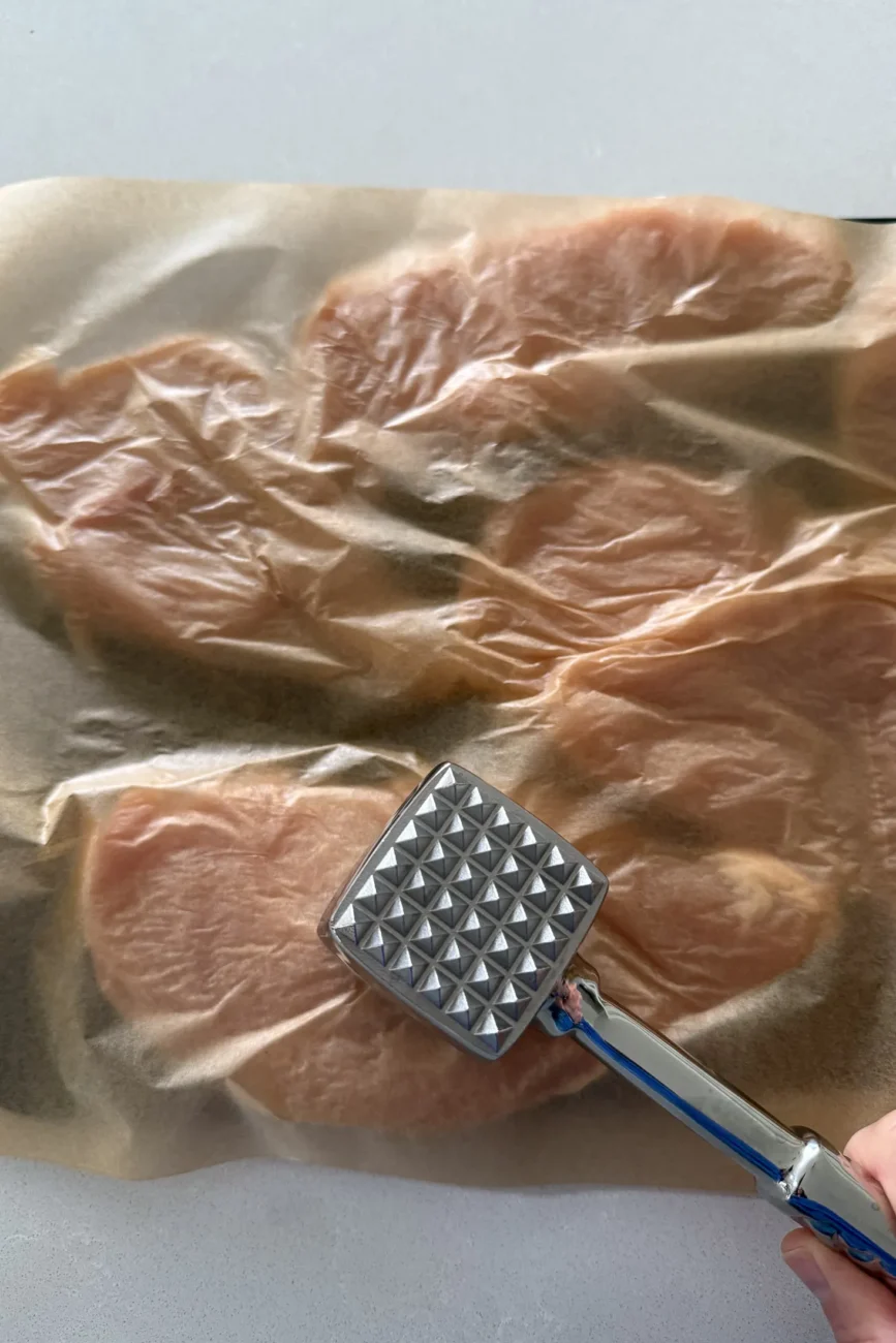 Flat side of meat mallet being used on chicken covered with baking paper